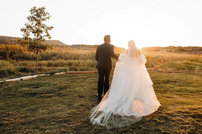 Bride with a flowing veil and groom walking in a sunset field at Sabrina Cedars in Abilene Texas, photographed by Double Knot P