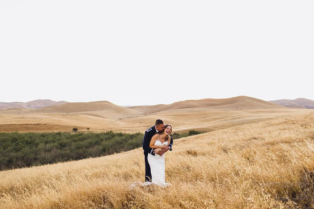 Bride and groom embracing in a golden field with hills in the background in California, photographed by Double Knot Photography, showcasing a tender wedding moment.