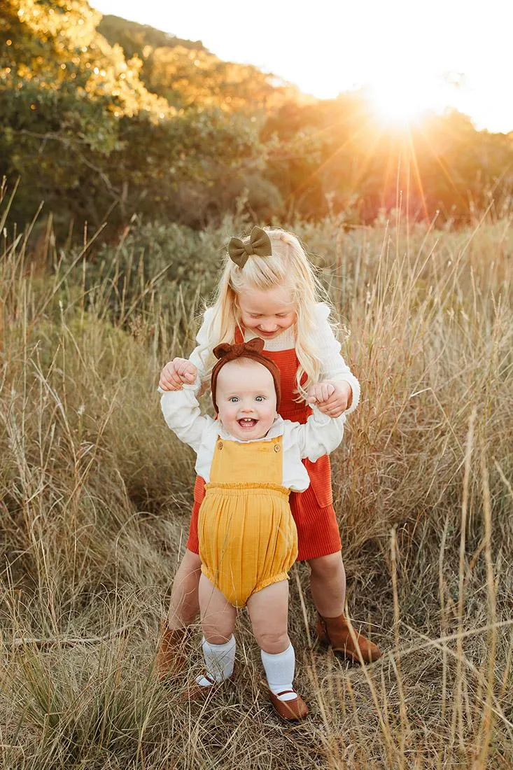 Two young siblings holding hands in a golden field at sunset, photographed by Double Knot Photography, showcasing a playful moment in a natural setting in Abilene Texas