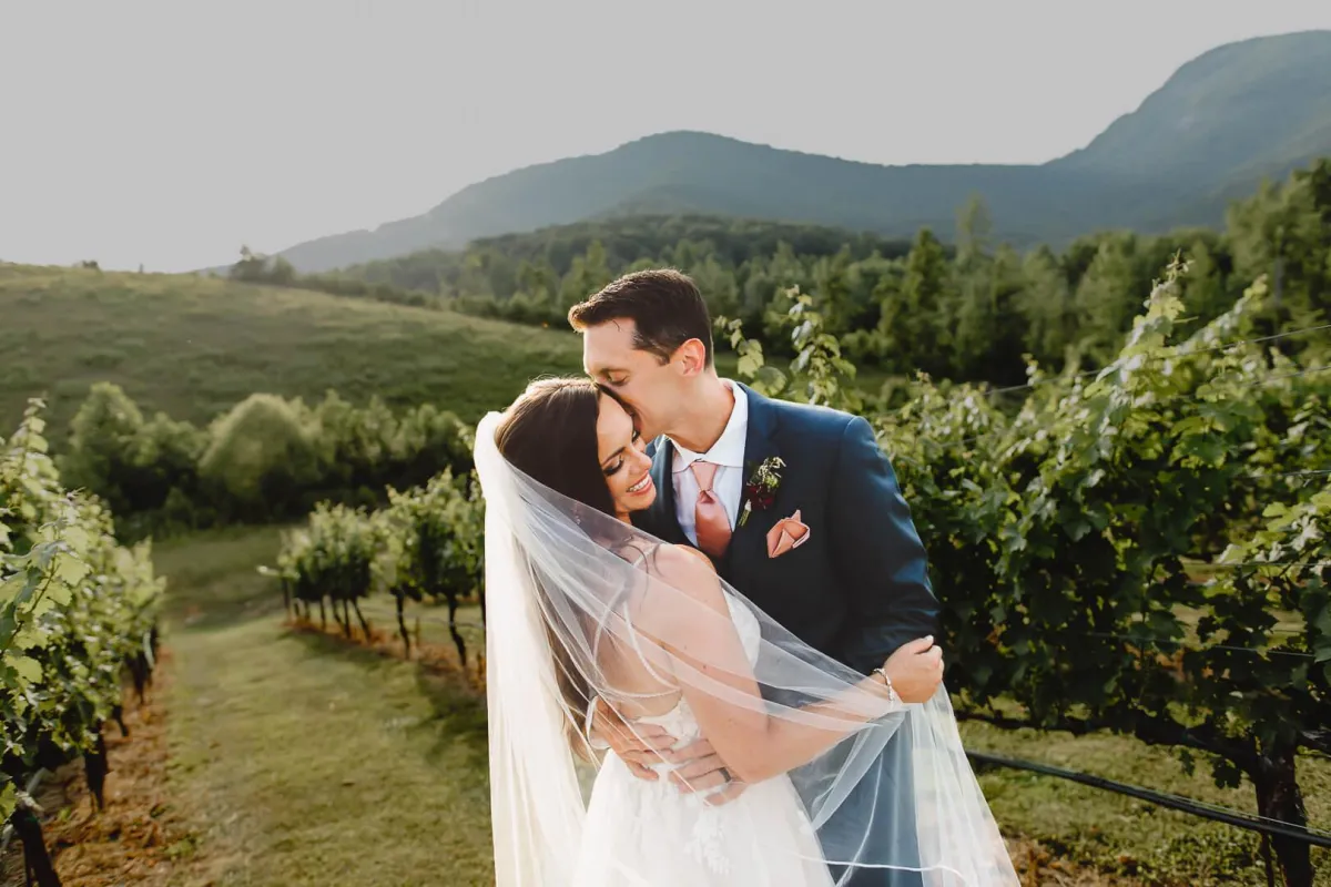 Bride and groom embracing in a vineyard with a mountain view, photographed by Double Knot Photography, highlighting a romantic wedding moment at Yonah Mountain vineyard