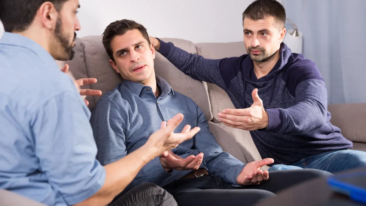 Four adults in a tense family meeting around a dining table reviewing will paperwork during an inheritance dispute, with scattered documents and an advisor present.