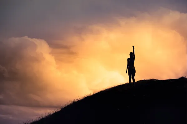 Woman standing on hilltop, with arm in air representing victory and freedom in setting healthy boundaries.