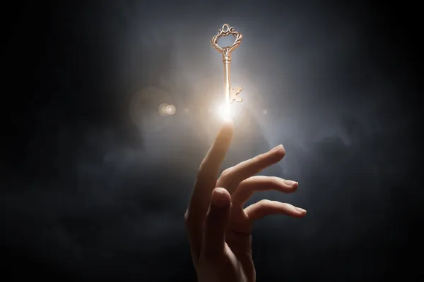 A hand with pointer finger reaching up fromt he darkness towards a key that is illuminated, representing transformation