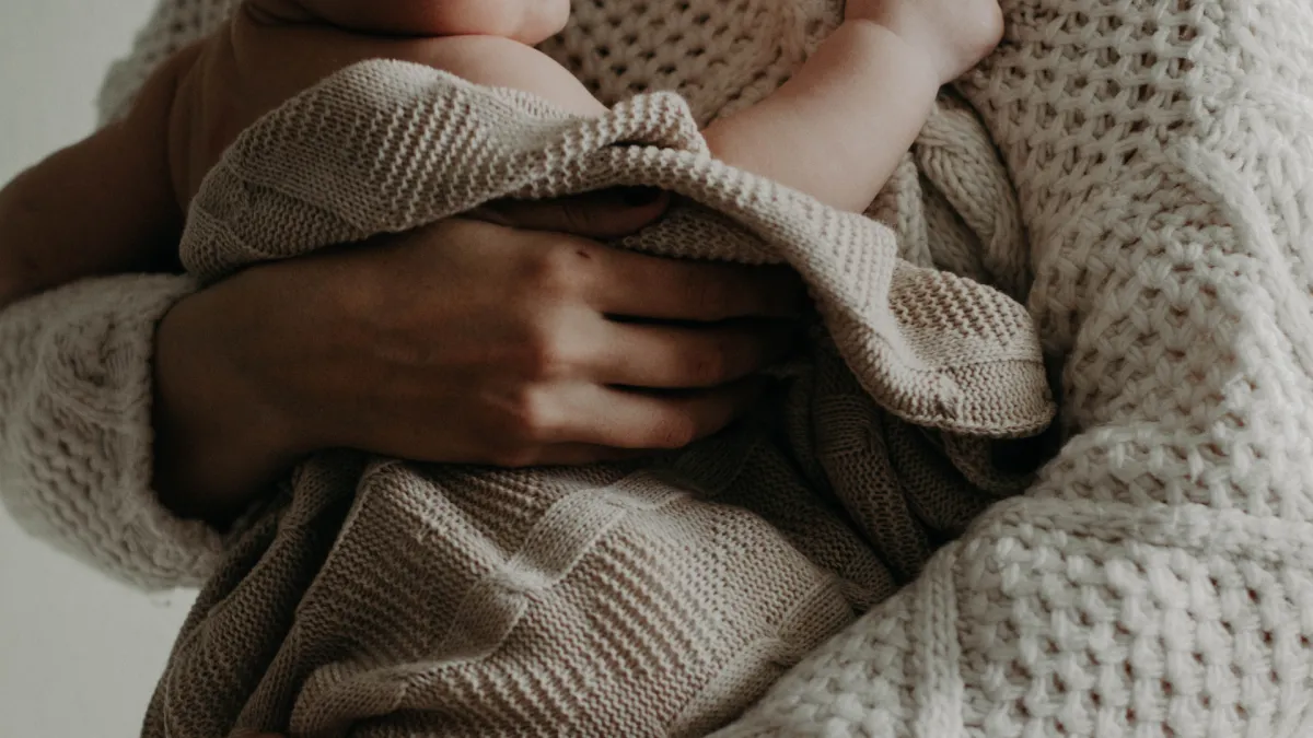 Close-up of a parent holding a newborn wrapped in a soft knit blanket, both dressed in cozy neutral-toned sweaters, conveying warmth and tenderness.