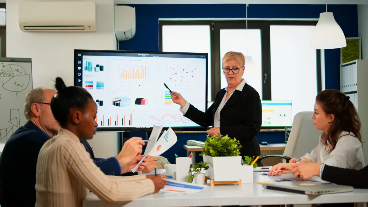 Business analyst presenting financial charts and data on a digital screen during a team meeting with diverse colleagues.