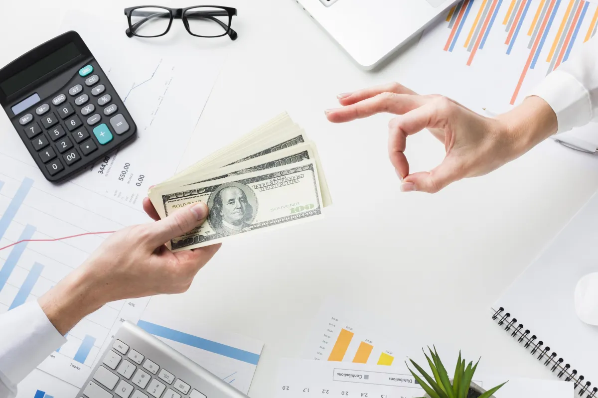 Businessperson handing over US dollar bills during a financial transaction at an office desk with charts and calculator.