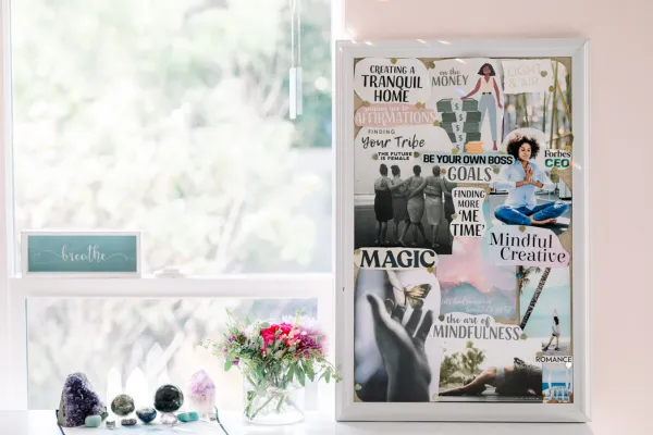 Vision board displayed in white frame on blush pink wall, featuring collaged images and words about mindfulness, creativity, goals, and home life. Windowsill below holds crystals, healing stones, and fresh pink flowers in a vase, with soft natural light filtering through.