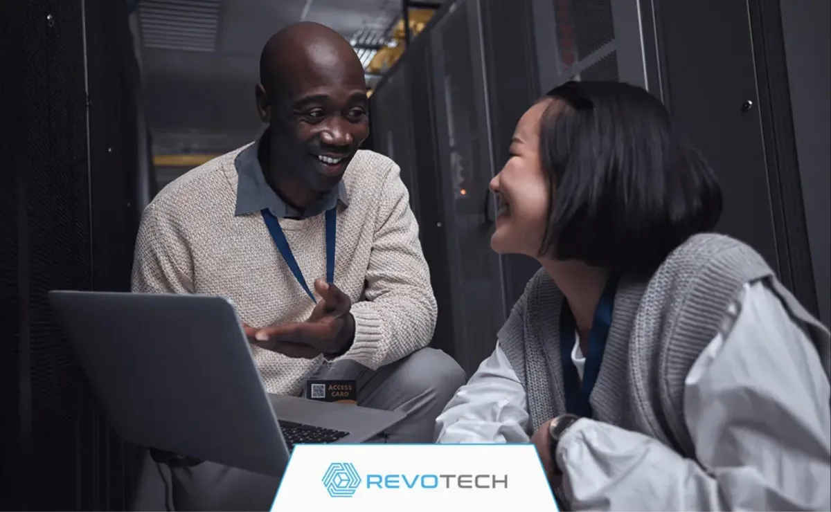 Two smiling IT professionals crouched by hardware and an open laptop discussing risk reduction.