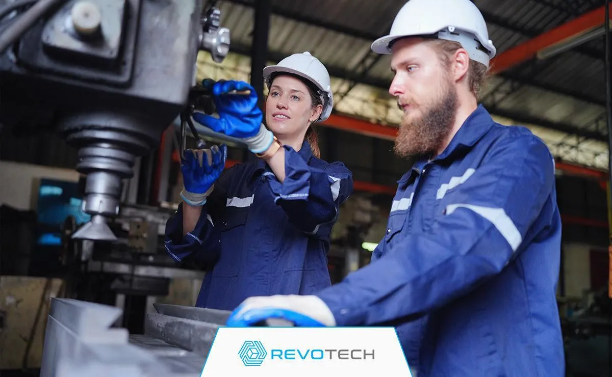 Image of two manufacturing workers in white hard hats and blue coveralls operating a machine at a manufacturing plant.