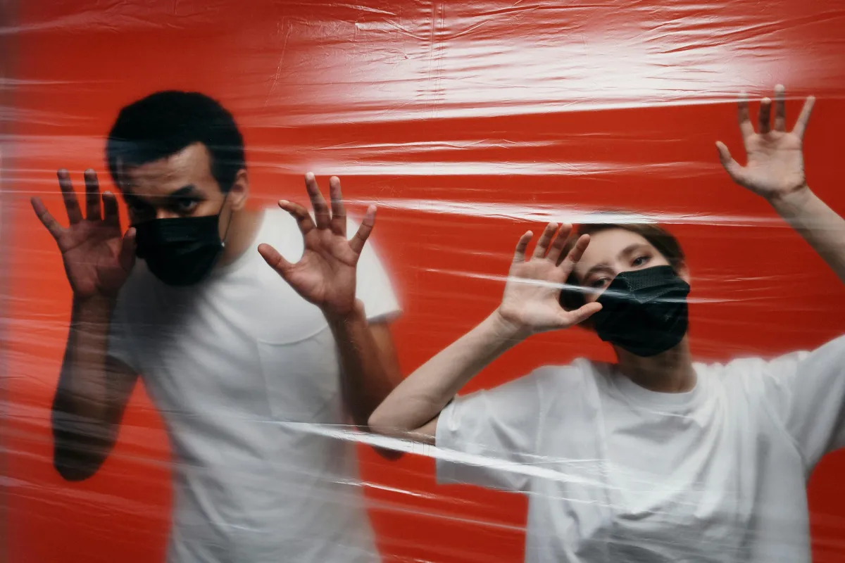 Two people wearing black masks stand behind clear plastic against a red background, symbolising the feeling of being trapped by intrusive thoughts and compulsive behaviours in OCD.