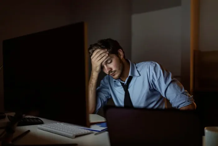 A tired man in a blue shirt sits at a desk late at night, leaning forward with his hand on his forehead while looking at a computer screen, conveying stress or exhaustion in a dimly lit office.