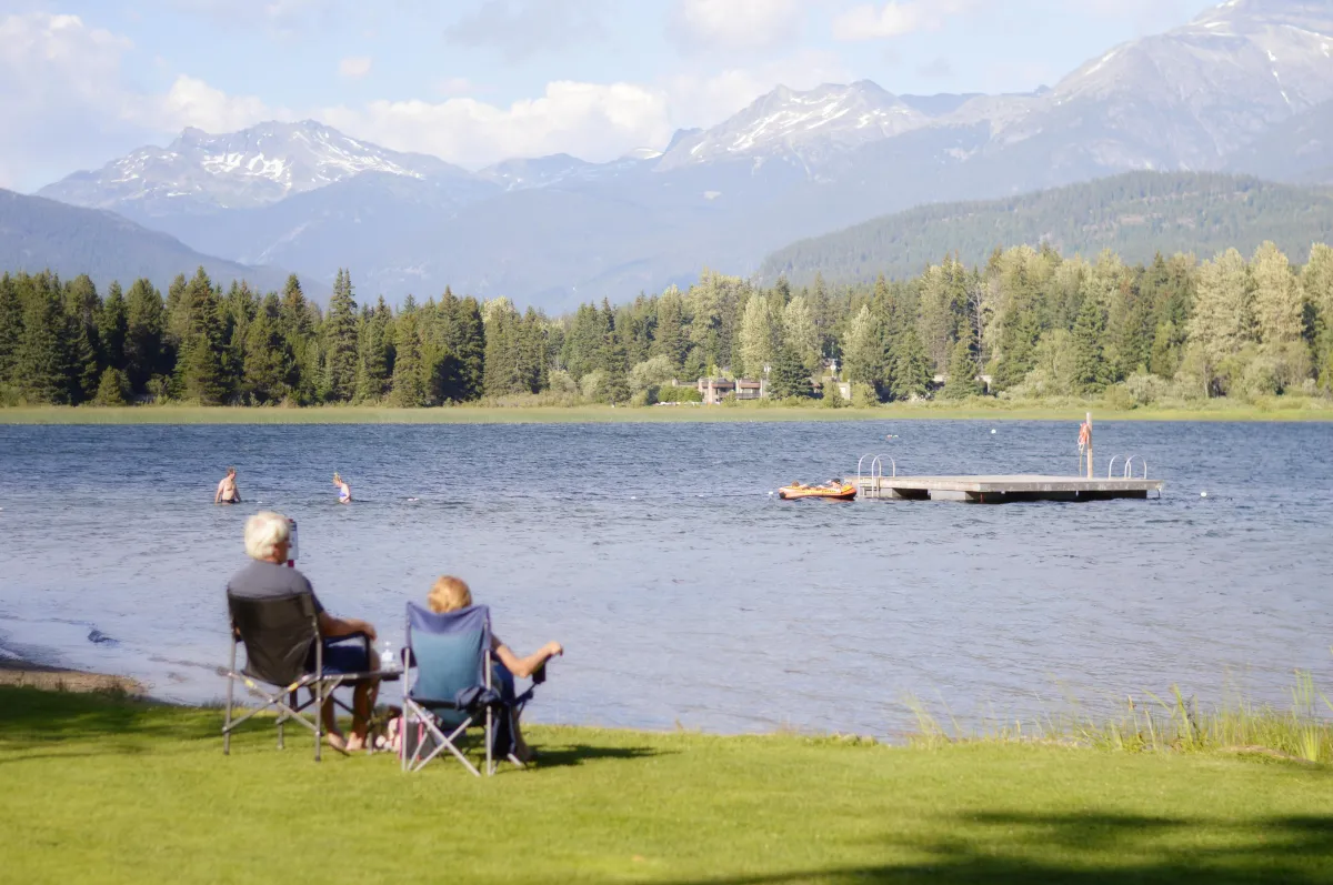 2 people sitting in chairs at the edge of a lake with trees and mountains in the distance