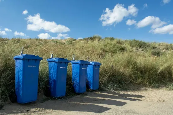 Row of blue trash bins placed near sand dunes under a bright blue sky with scattered clouds