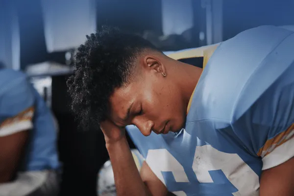 Exhausted young football player sitting alone on sideline with head down showing signs of athlete burnout and mental fatigue
