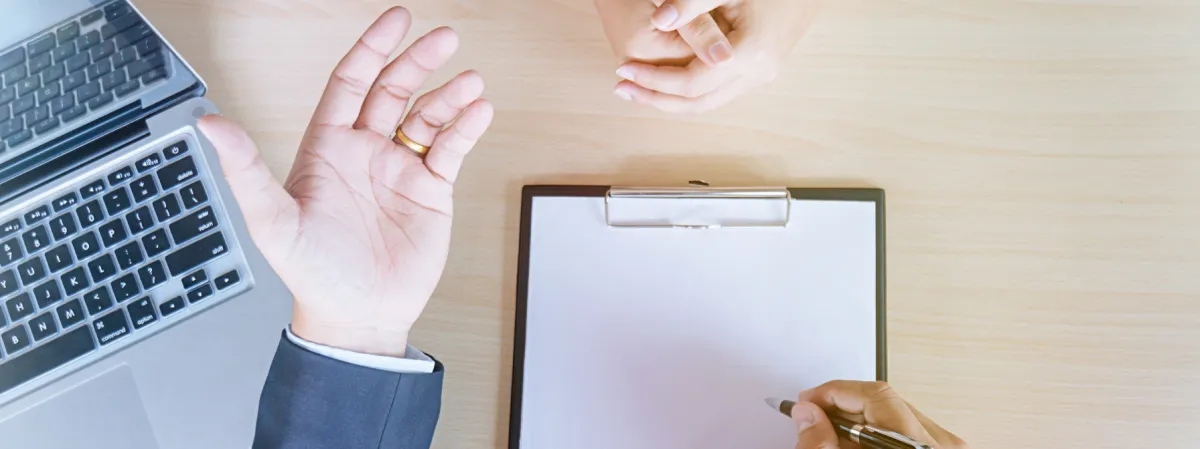 A close-up of hands on a wooden desk, with one gesturing, another holding a pen, a clipboard with blank paper, and part of a laptop keyboard visible.