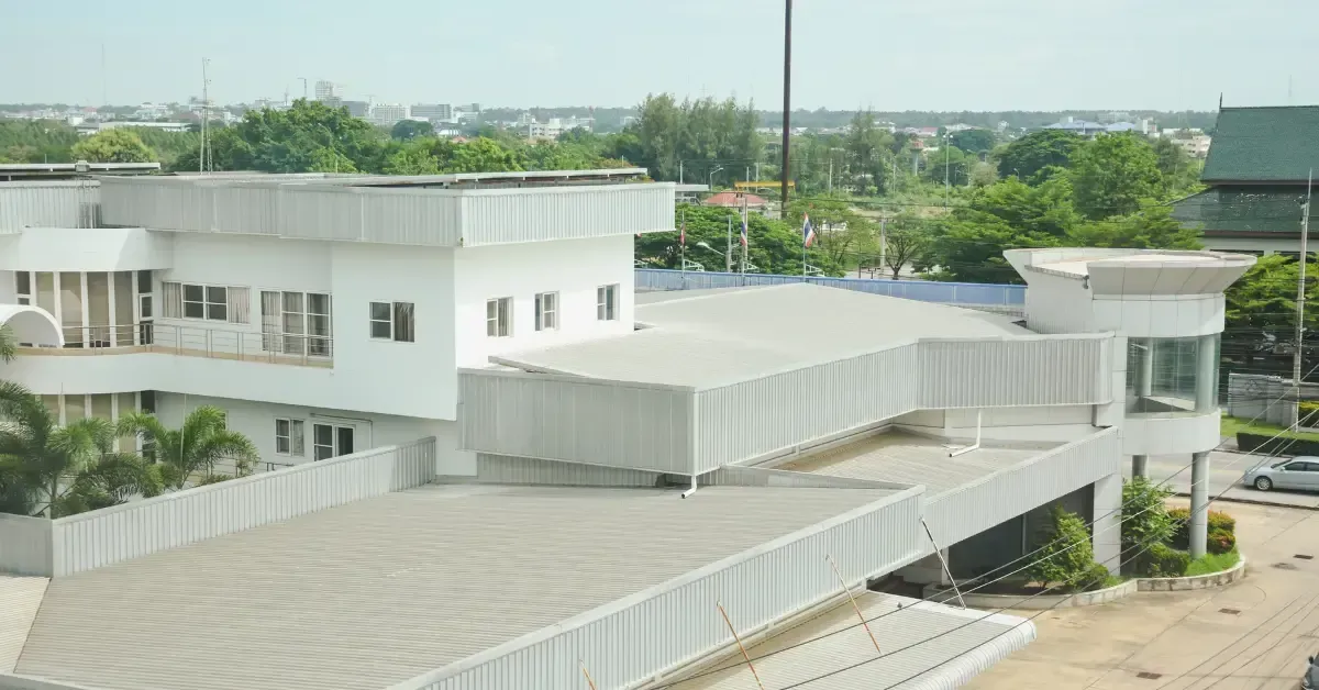 Modern commercial building with flat, light-colored metal roofing, surrounded by greenery and situated in an urban area.