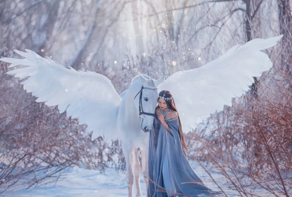 A woman in a long blue dress is close to a white Pegasus, both are standing in the snow.
