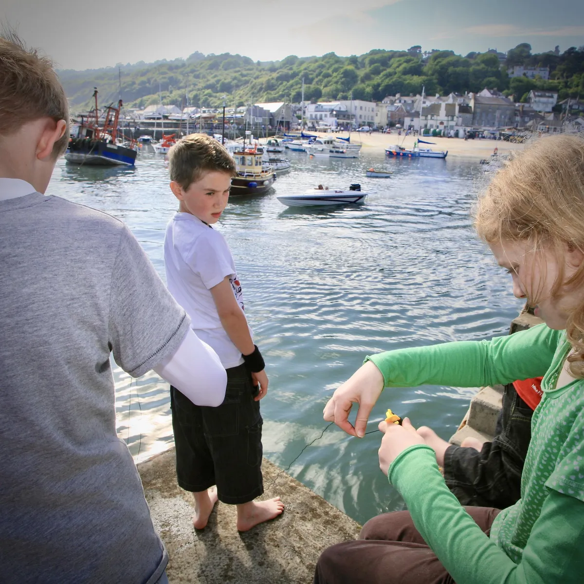 Children crabbing off the harbour wall in Lyme Regis with colourful buckets, a view of the Cobb with fishing and day boats behind.