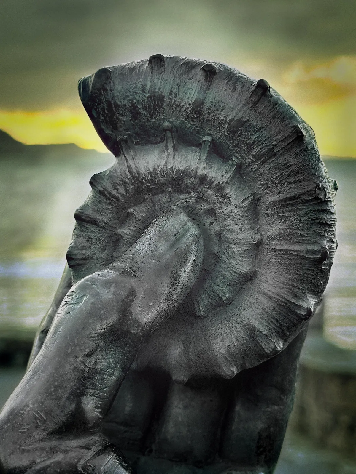 Close-up of a bronze sculpture of a hand holding an ammonite fossil, photographed against the dramatic Dorset coastline at sunset, symbolising fossil hunting on the Jurassic Coast.