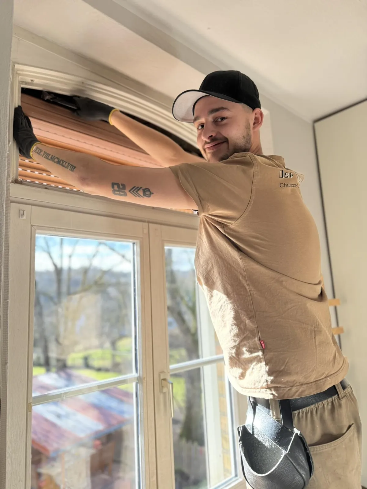"Handwerker in Arbeitskleidung und schwarzer Kappe repariert oder installiert einen Rollladen an einem Fenster mit Rundbogen."