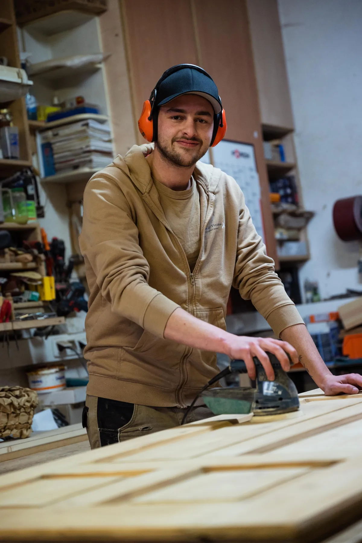Ein junger Tischler mit Gehörschutz und schwarzer Kappe schleift eine Holztür in einer Werkstatt.