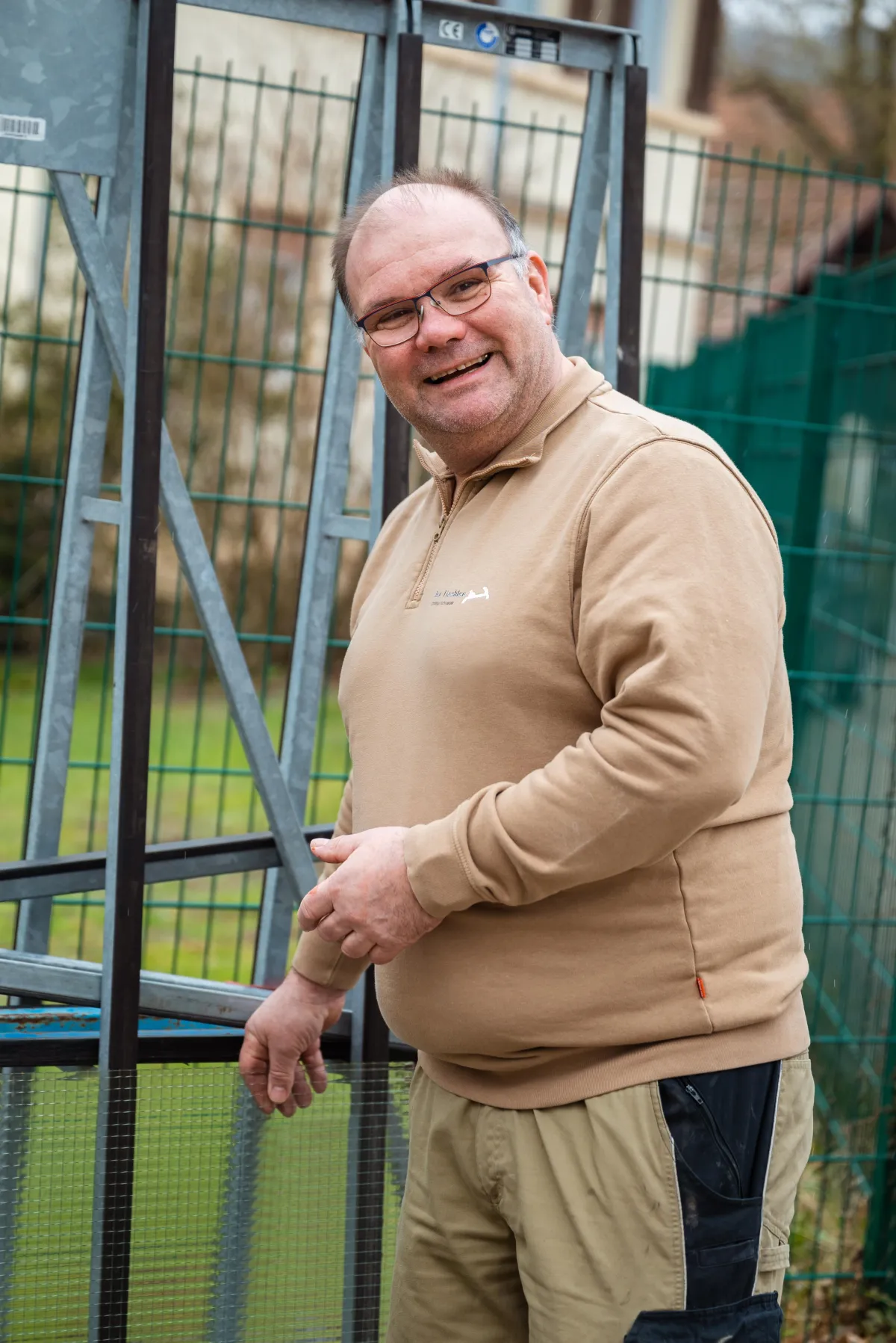 Handwerker mit Brille und beigem Pullover steht lächelnd vor einem Metallzaun und blickt in die Kamera