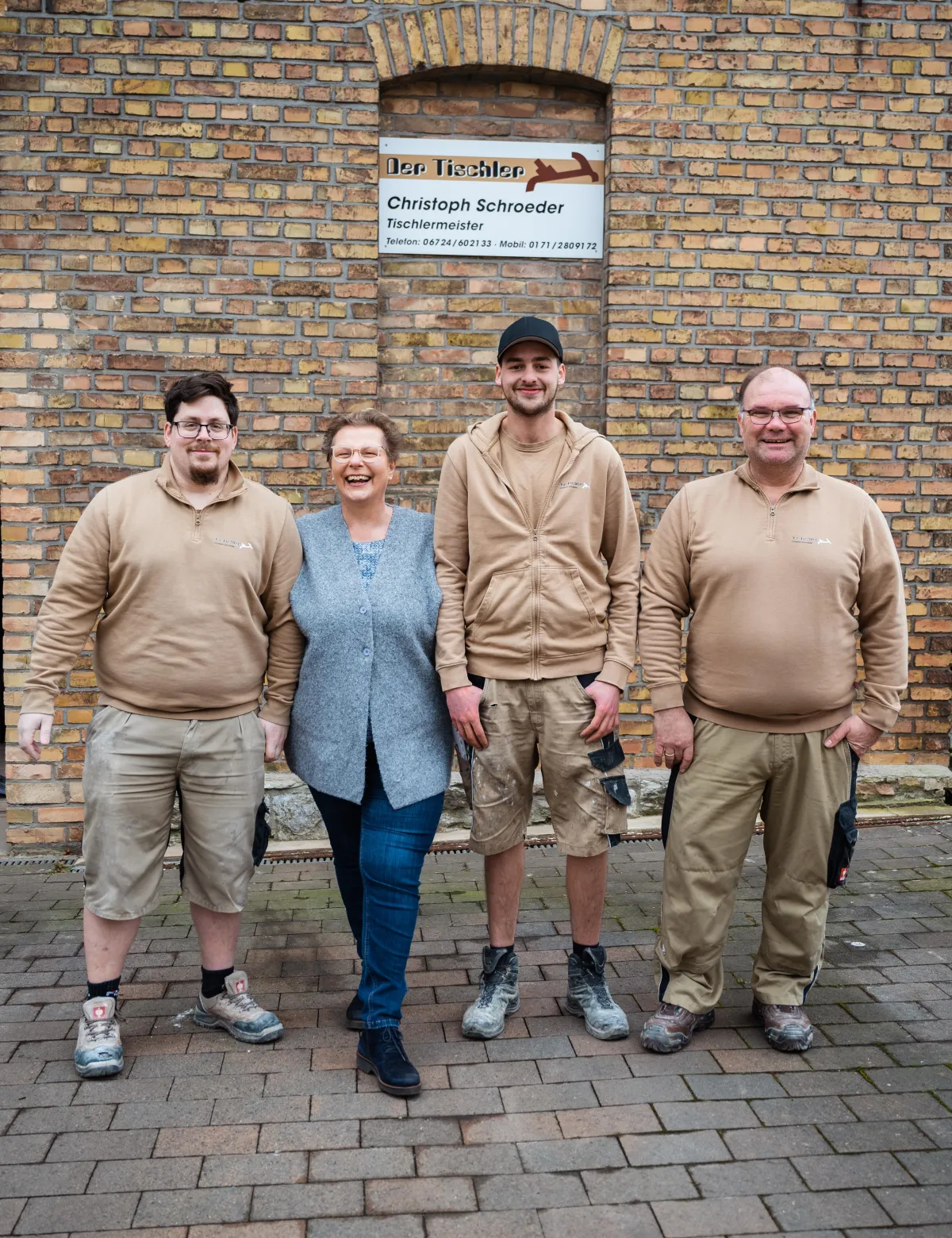 Vier Personen stehen vor einer Backsteinwand mit einem Schild der Tischlerei 'Der Tischler Christoph Schroeder' und lächeln in die Kamera.