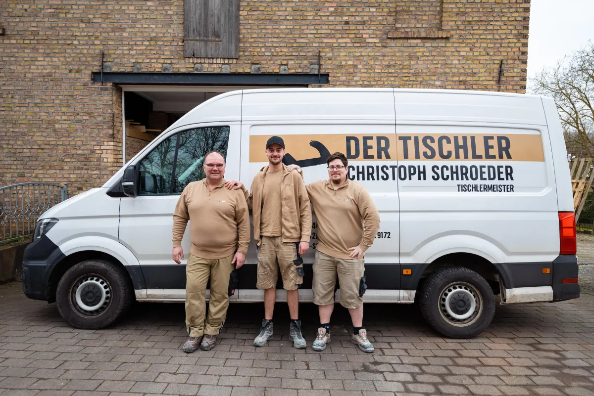 Drei Handwerker in beiger Arbeitskleidung stehen vor einem weißen Firmenwagen mit der Aufschrift 'Der Tischler Christoph Schroeder' und lächeln in die Kamera.