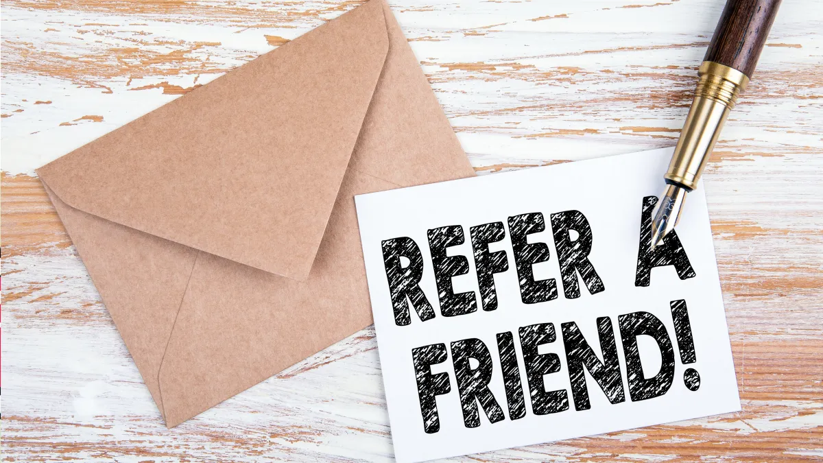 Card reading "REFER A FRIEND!" beside a kraft envelope and fountain pen on a wooden table, referral program visual for small businesses using MyToolBox Pro CRM.
