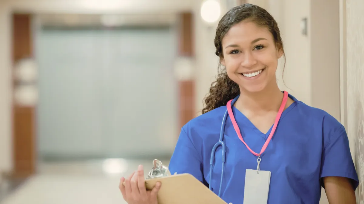 A confident young international nurse in blue scrubs smiles, ready to start her career after choosing the right state to take the NCLEX.