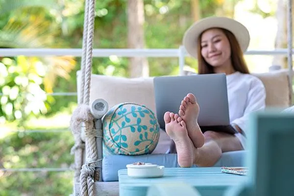 Female employee in a hat working remotely on a comfy sofa, with a sunny day in the background, symbolizing equitable coverage for remote and hybrid teams