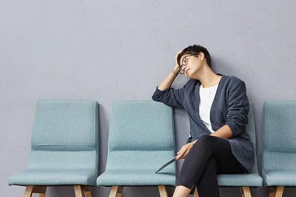 A woman sits on a row of chairs, resting her head on her hand while holding a document.