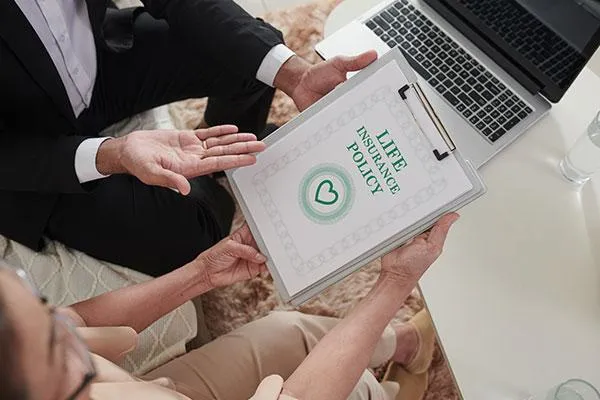 A person holds a clipboard with a life insurance policy document while discussing with another person