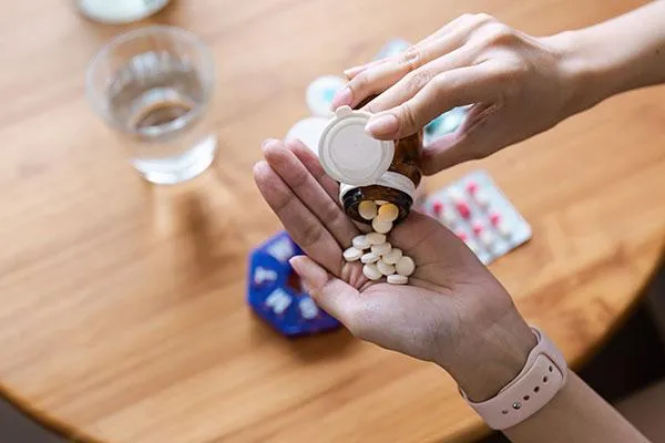 A person pours white pills from a bottle into their hand over a wooden table with medication and a glass of water