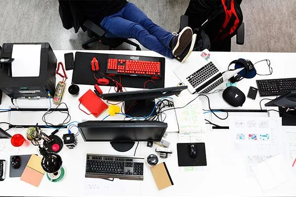 A cluttered office desk with multiple computers, gadgets, and office supplies, with a person resting their feet on a chair