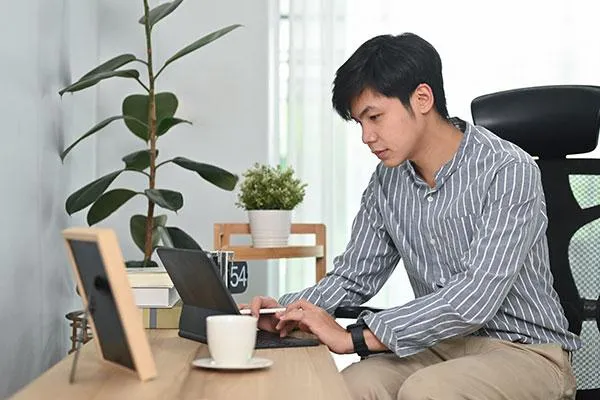 A young man working on a laptop at a desk with plants, a coffee cup, and framed photos