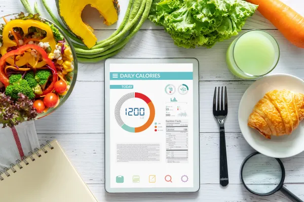 Tablet displaying daily calorie tracking surrounded by fresh vegetables, fruit, and breakfast foods on a white table