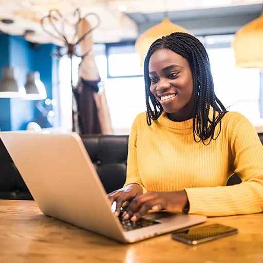A woman sitting in a cafe, looking at her laptop with her phone next to her