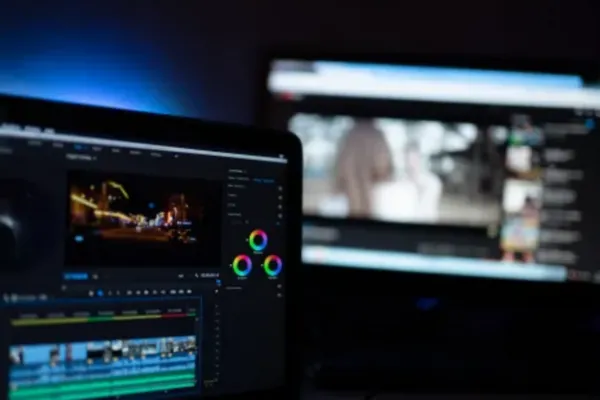 A person is using a computer with video editing software, featuring various video clips and color adjustments. Another screen in the background displays a blurred video player interface.