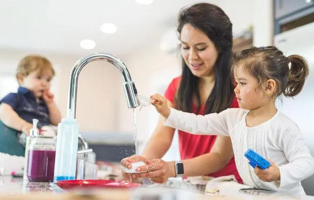 Mother helping her young daughter wash dishes at the kitchen sink, illustrating the hands-on responsibilities of a stay-at-home parent, while a toddler sits nearby, emphasizing the multifaceted role of caregiving and household management.