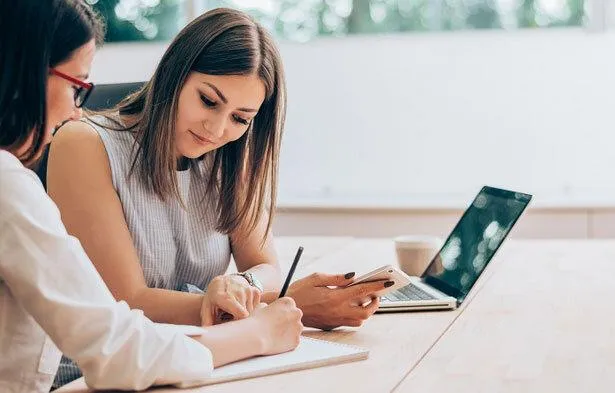 Two professional women discussing documents at a desk, with a laptop and notepad, symbolizing legal consultations or career planning support, relevant to topics like alimony factors or advancing a spouse’s career.