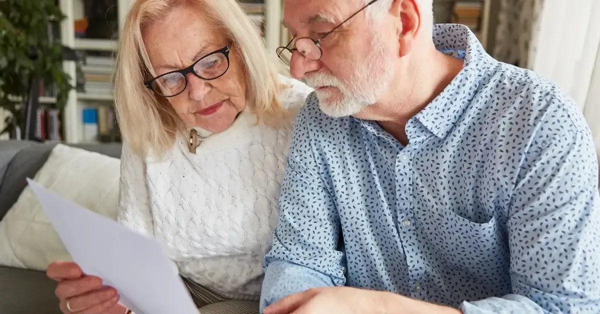 An elderly couple wearing glasses sitting together on a couch, closely reading a document.