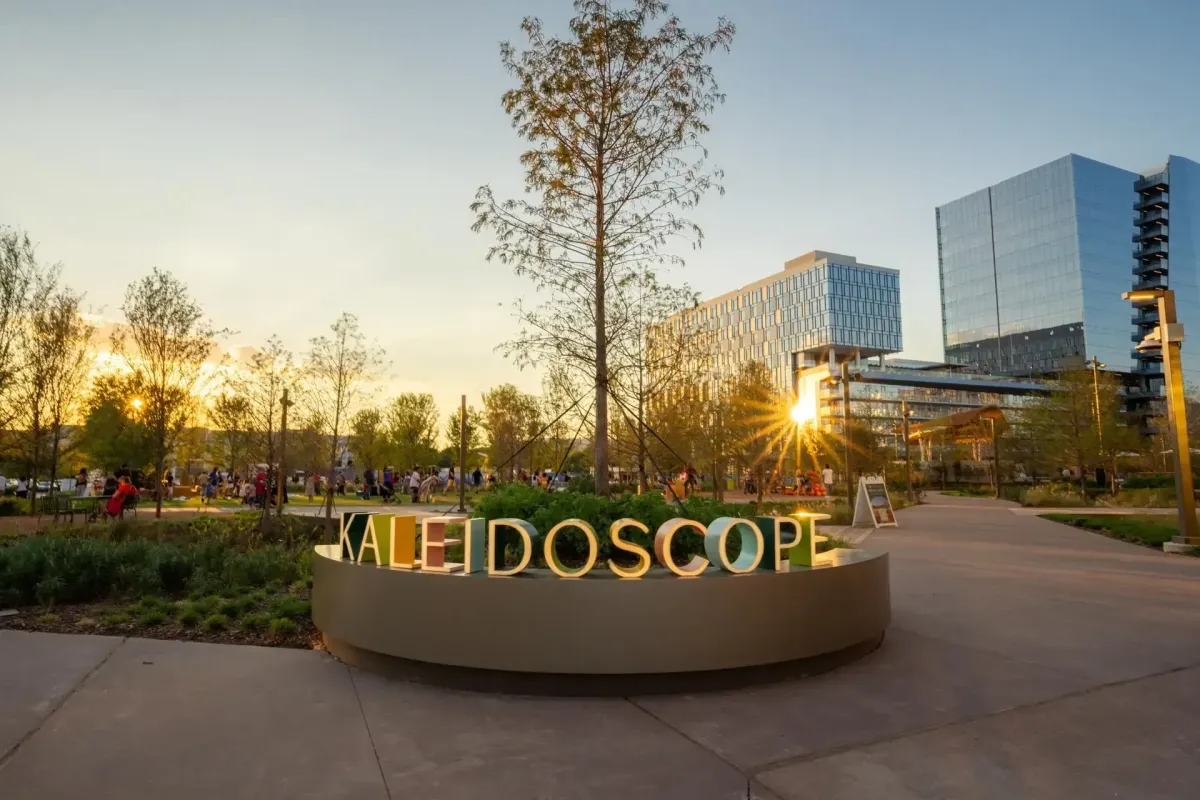 Modern park with glowing “Kaleidoscope” sign, people gathering at sunset, and glass buildings.