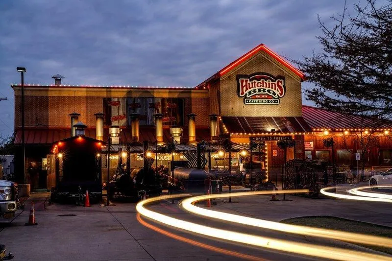 Hutchins BBQ lit up warmly with smokehouse pits and evening car light trails.