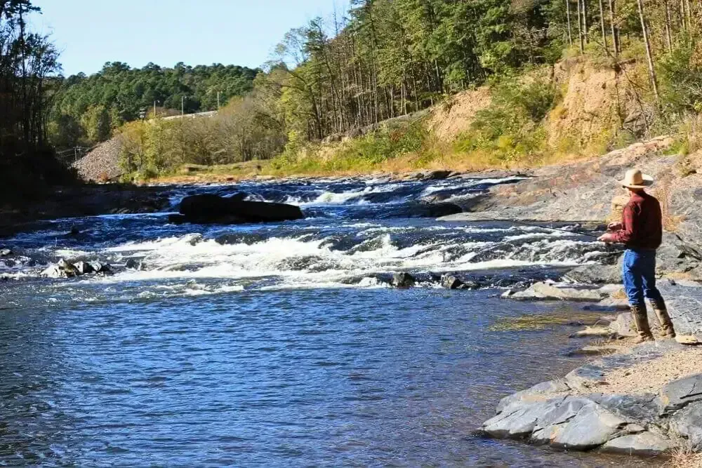 Man fishing on rocky riverbank with forested hills and flowing water.