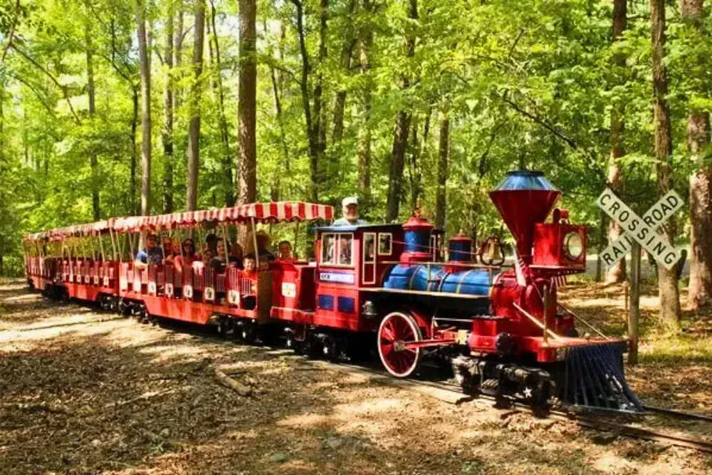 Miniature red train in a lush forest, passing a quaint railroad crossing sign.