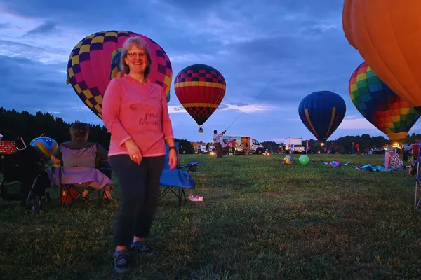 Woman smiling in front of colorful hot air balloons at a festival during sunset, wearing a pink sweatshirt with the words ‘just breathe.’ Several balloons are inflating in the background against a dramatic evening sky.