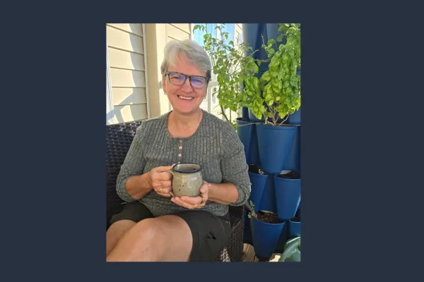 Claudette smiling on porch with coffee mug in hand, sitting beside vertical herb garden filled with basil