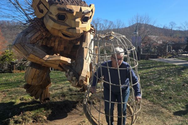 A mature woman stepping out of a rope-style cage beside a large wooden troll sculpture on a sunny trail, symbolizing lighthearted adventure and personal growth.