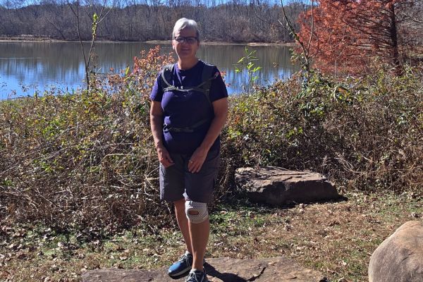 Woman standing on a nature trail by a lake during a calm morning hike, representing rebuilding strength and steady movement in her 60s.